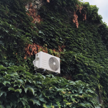 External white air conditioner unit on the wall among green leaves of wild grapes. Private house and modern equipment for microclimate.