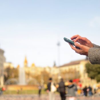 Woman hands holding a smartphone and typing message, connecting online in a blurred city square with people and a historic building in barcelona, catalonia background