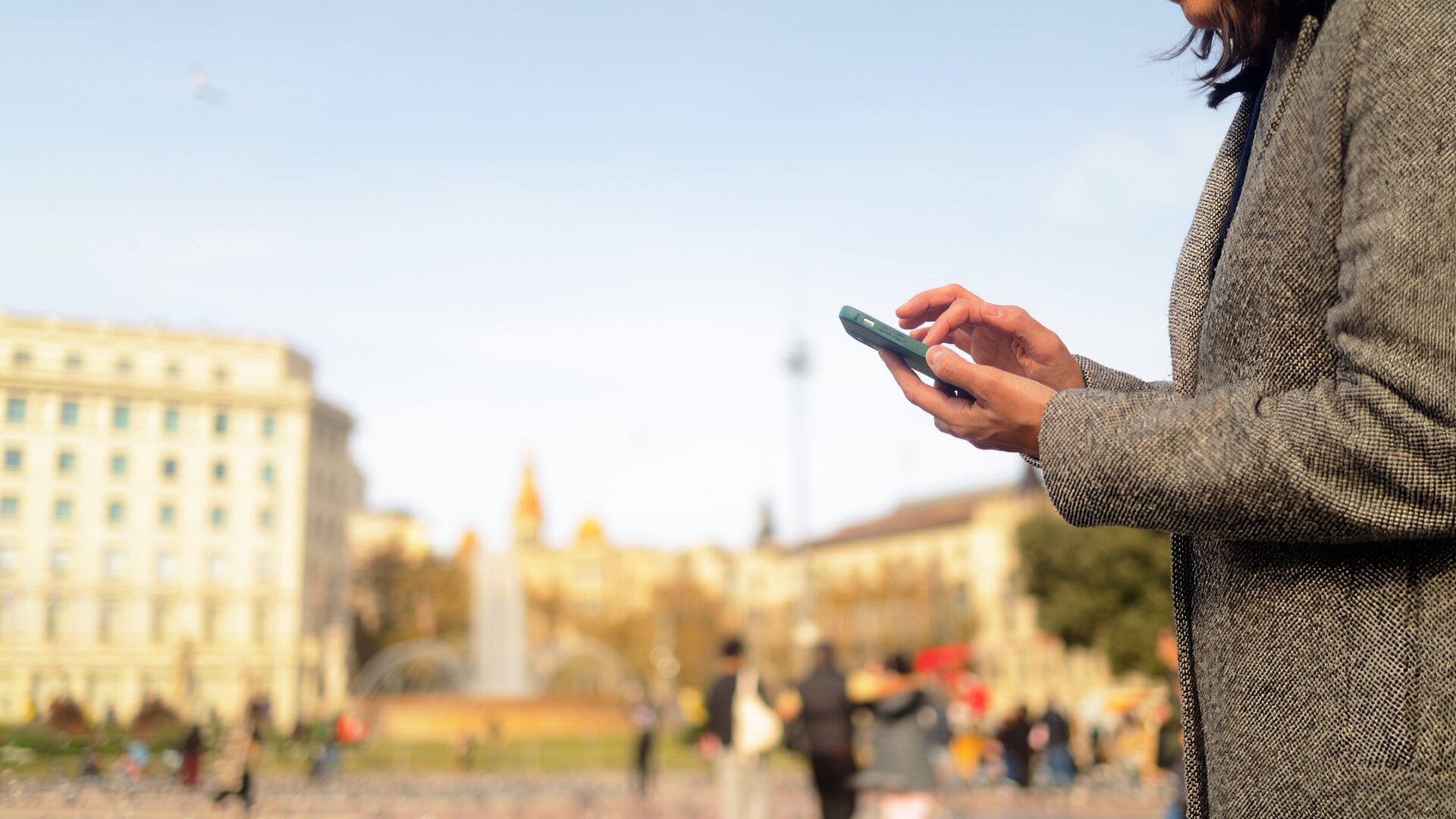 Woman hands holding a smartphone and typing message, connecting online in a blurred city square with people and a historic building in barcelona, catalonia background