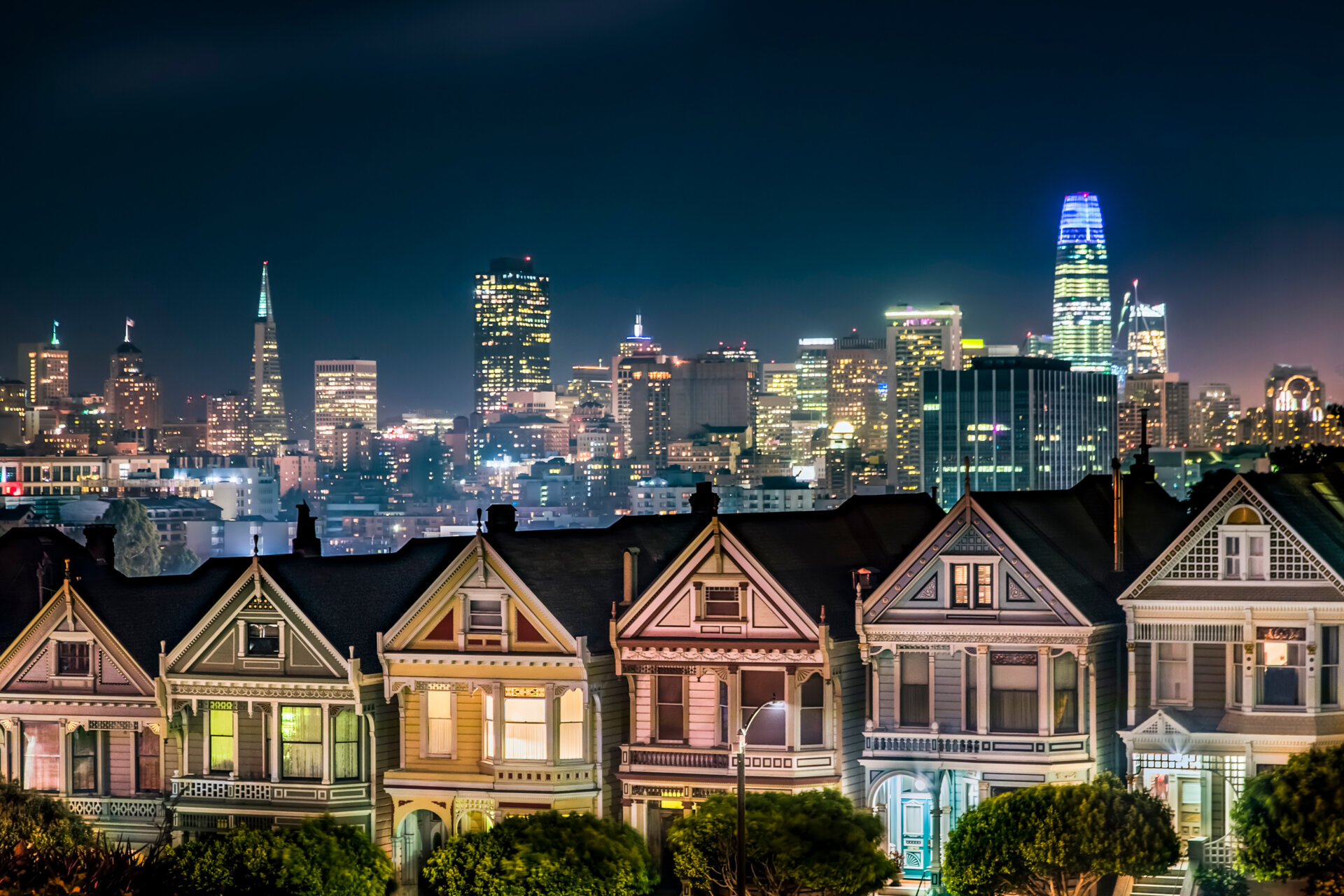 Backed by the night skyline of the city of San Francisco, California, the Victorian era houses near Alamo Square Park, are painted in colors to accentuate their architectural details.