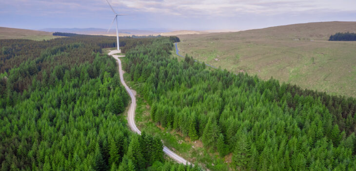 Aerial view on a wind farm turbine build in a rural country area with green forest. Making power using power of nature. Blending technology into nature environment. Cloudy sky. Electricity production