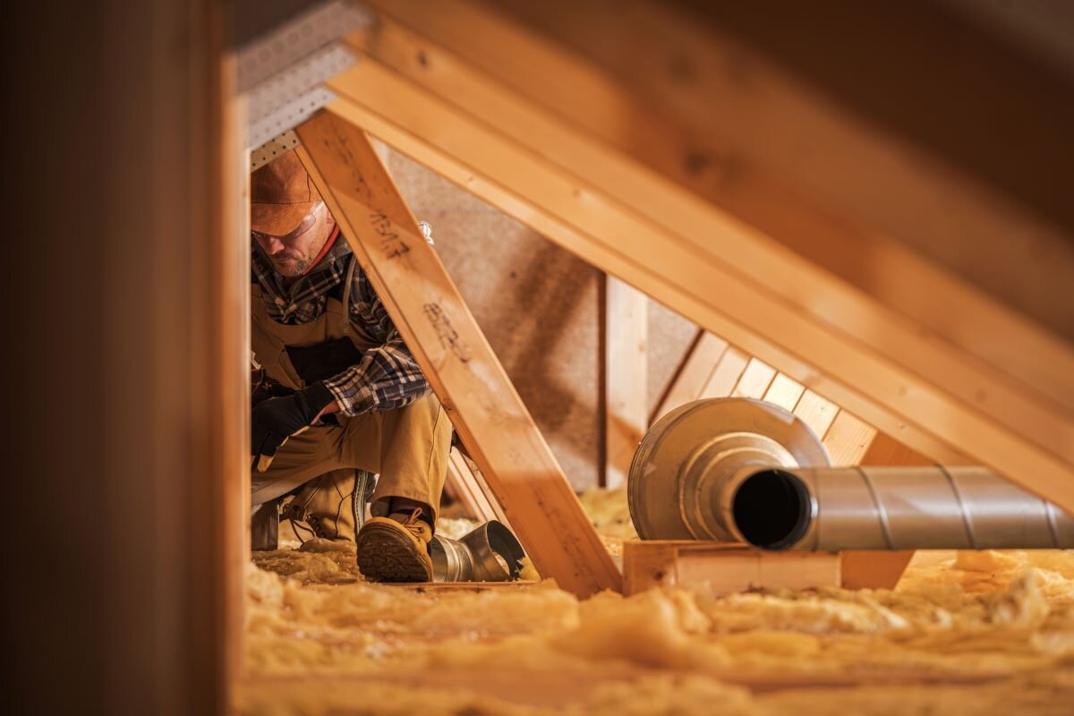 Air ventilation installer working in attic. He is wearing safety equipment and work overalls, and is crouched under wooden beams.