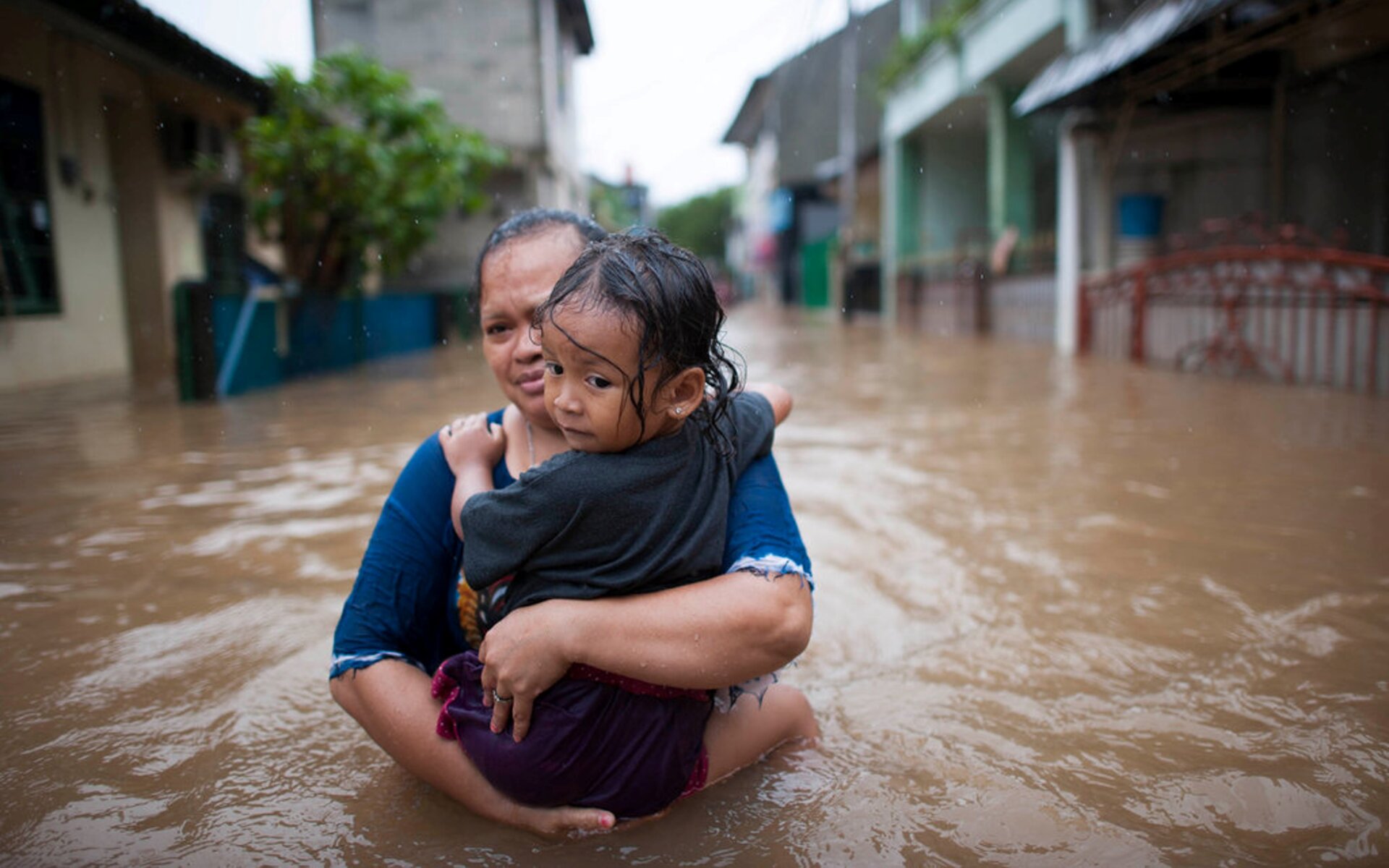 The image shows a woman carrying a child as they wolk through a flooded street, showcasing the vulnerability to climate risk, loss and damage.