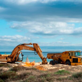 Digger and truck on a beach moving sand in a beach, with a blue sea and a blue sky in the background.