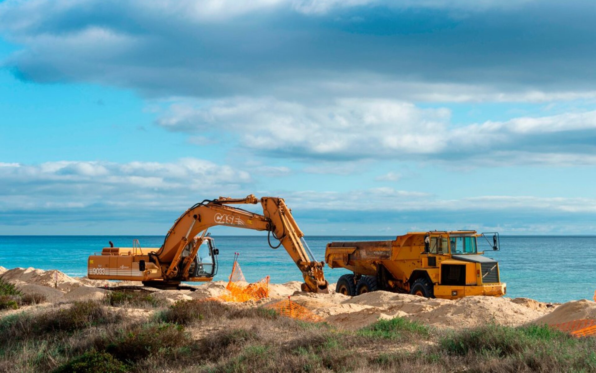 Digger and truck on a beach moving sand in a beach, with a blue sea and a blue sky in the background.