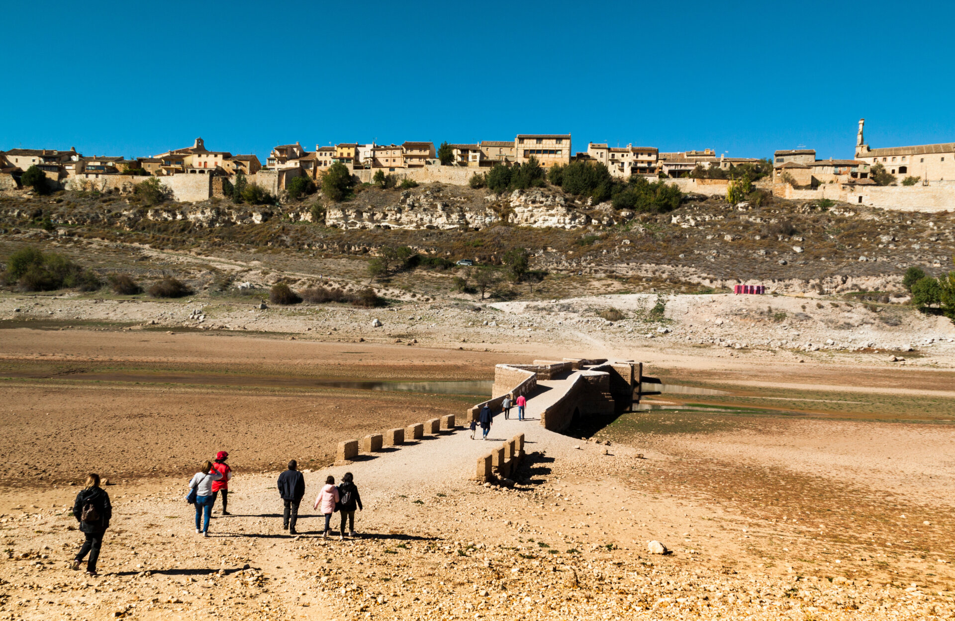 The image shows a group of people crossing a bridge over a dry river in Segovia, Spain. In the background, there is a dry mountain with a small village on the top.