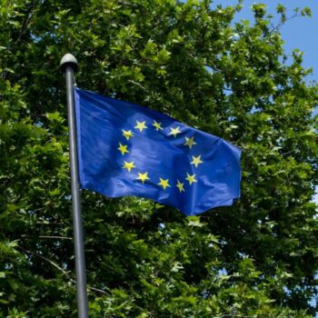 EU flag waving with the green leaves of a tree in the background. Photo via Adobe.