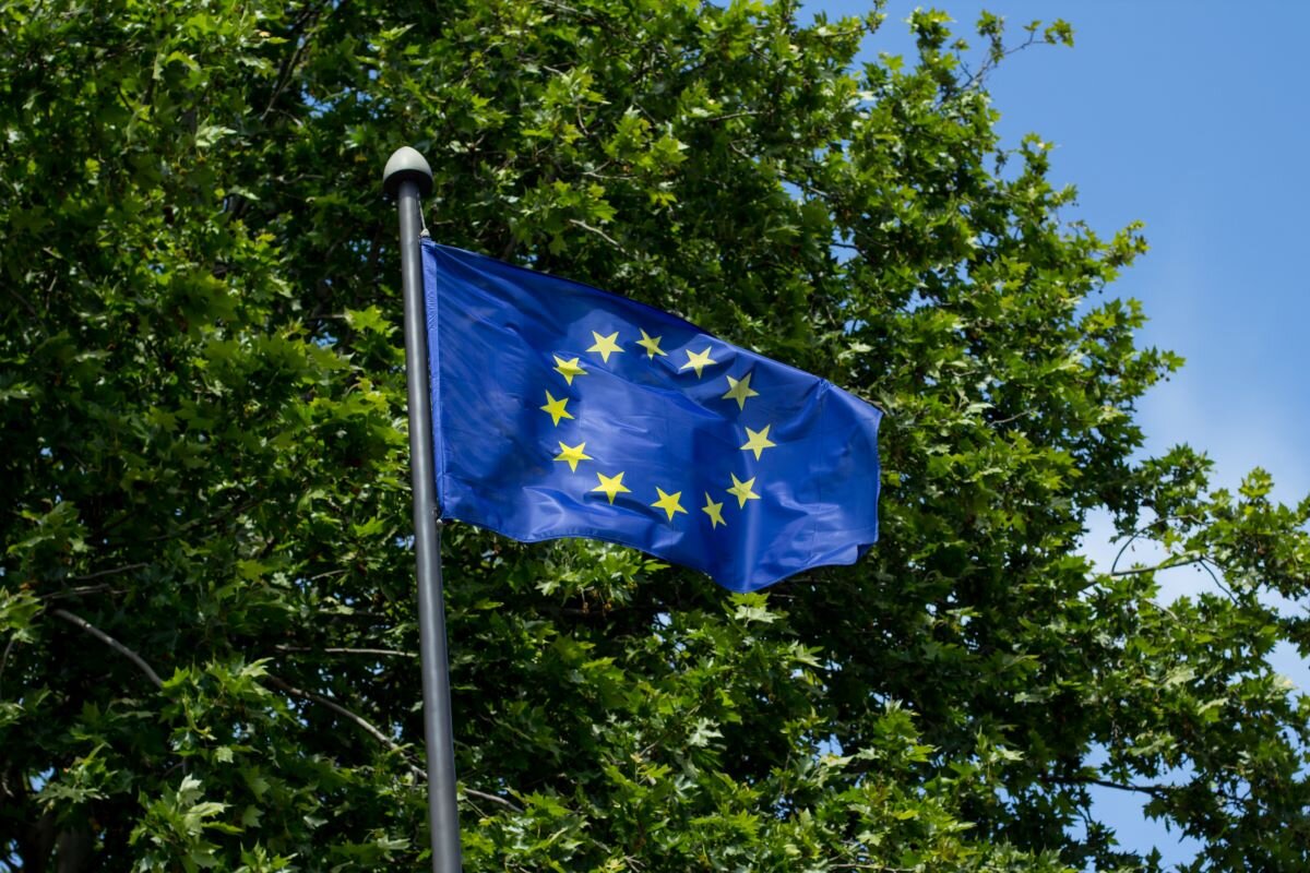 EU flag waving with the green leaves of a tree in the background. Photo via Adobe.