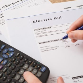 The image shows a calculator on several energy bills with prices together with a hand holding a blue pen.
