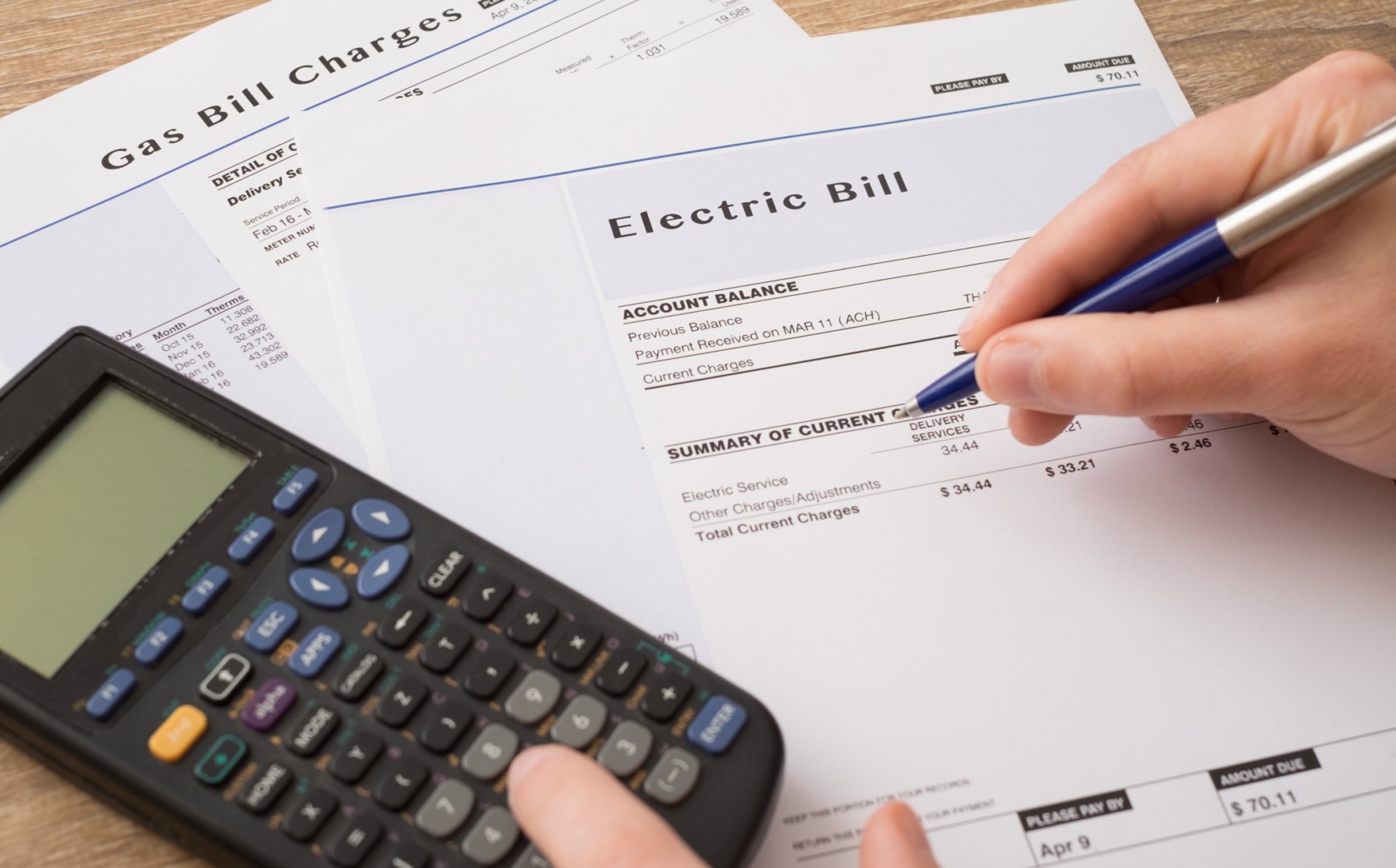 The image shows a calculator on several energy bills with prices together with a hand holding a blue pen.