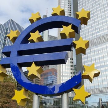 The image shows a sculpture with the shape of the Euro currency symbol in front of the European Central Bank headquarters in Frankfurt, Germany.