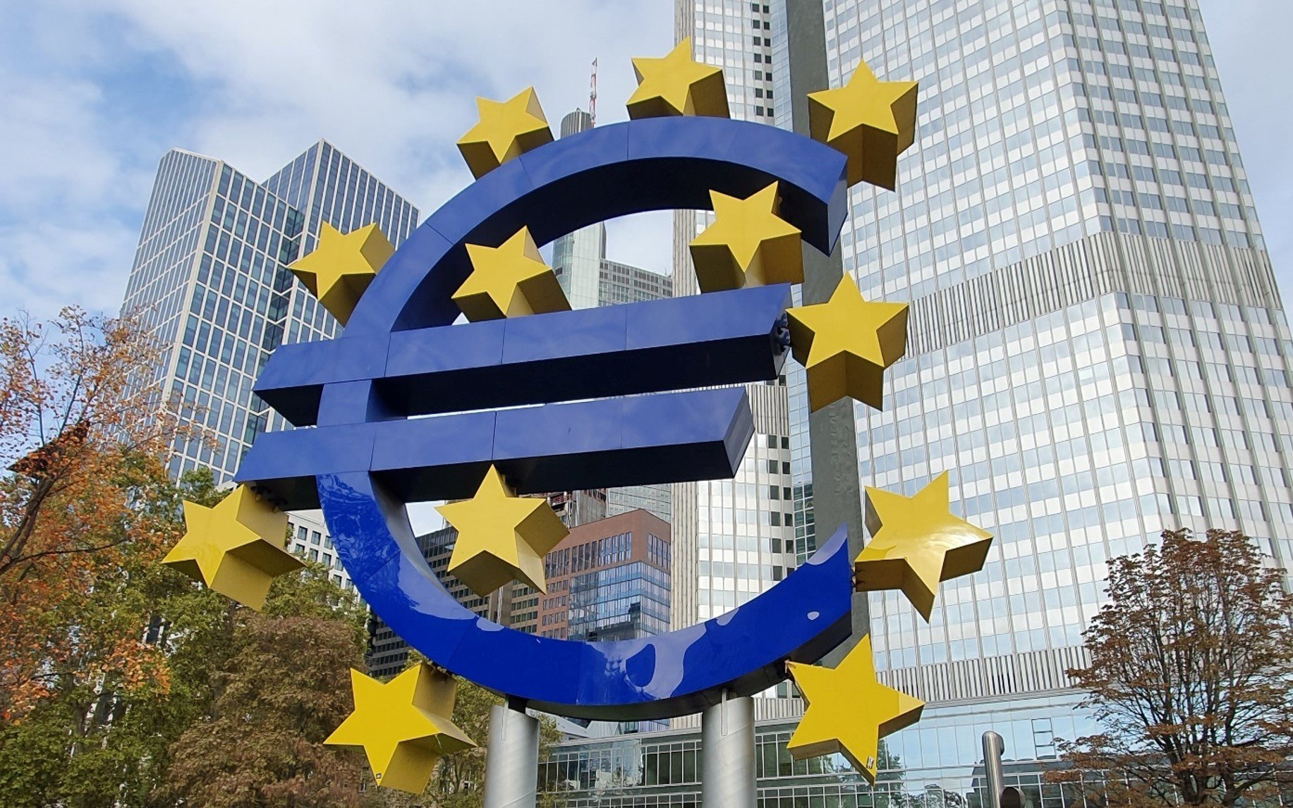 The image shows a sculpture with the shape of the Euro currency symbol in front of the European Central Bank headquarters in Frankfurt, Germany.