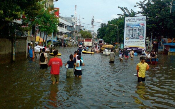 The image shows people walking in the water of a flooded marketplace, with a billboard on the background announcing discounts.