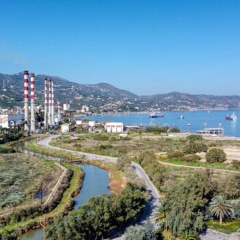 Industrial and trade area in Linoperamata, Greece. The image shows an industrial facility with chimneys over a green field, next to a river and the sea. There are cargo ships on the sea and mountains in the background.