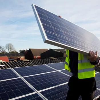 A builder holds a large solar panel in front of his face. Installing solar panels, a key part of energy security, on the roof of a home in Balcombe, UK.