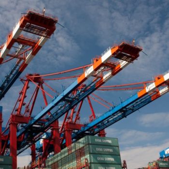 International shipping containers from China Shipping. Three cranes pick up containers against a blue sky.