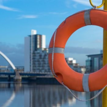 Orange circular life preserver in the foreground attached to a pole, in the background is the River Clyde in Glasgow, with a bridge and blue sky.