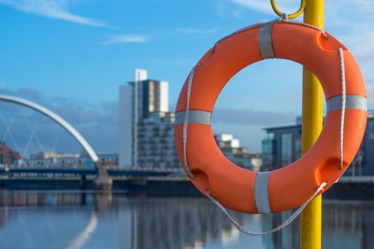 Orange circular life preserver in the foreground attached to a pole, in the background is the River Clyde in Glasgow, with a bridge and blue sky.