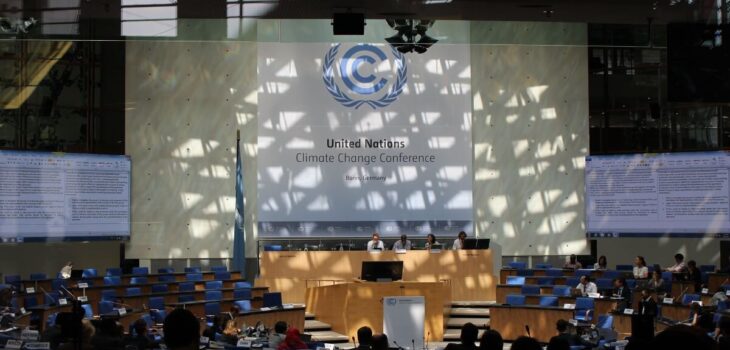 A sign for the UNFCCC in front of a room of people at desks. Negotiations commence in the SBs UNFCCC Plenary Room in Bonn, Germany, at the Bonn Climate Conference in 2015.