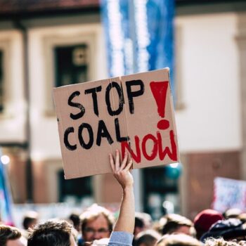 The image shows a hand holding a placard which reads "Stop coal now!". On the lower part of the image, you can see many heads of people as in a demonstration.