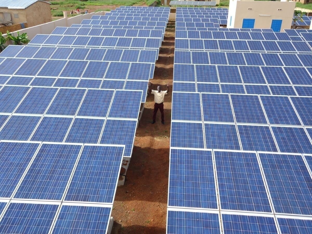 Man stands in a gap between blue solar panels, aerial view. Solar energy farm in Kolondiéba, Mali provides an alternative to thermal diesel to provide electricity. Photo by CIF Action on Flickr.