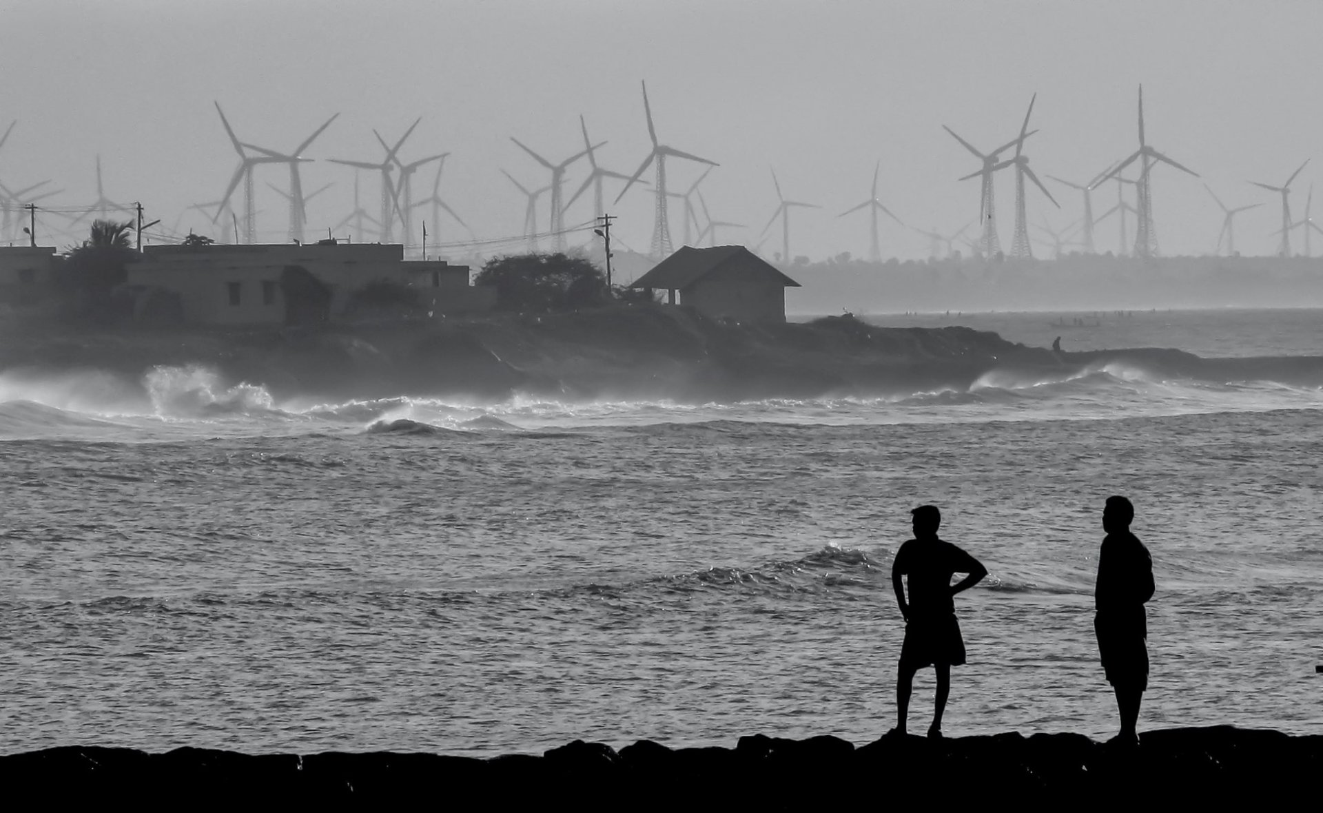 Wind turbines and two people standing by the ocean in Kanyakumari, Tamil Nadu, India. Black and white photo