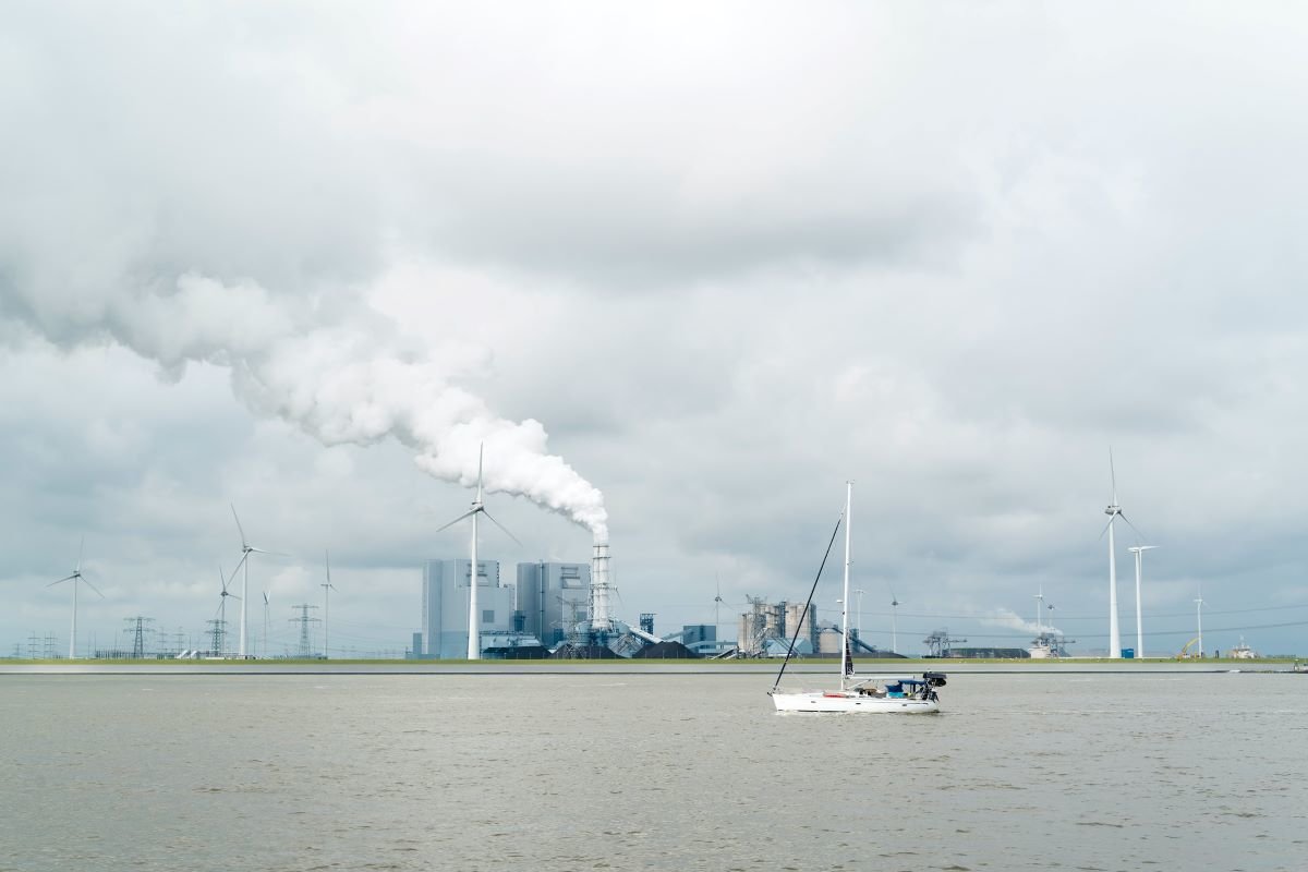 Wind turbines set against industrial smoke in Borkum, Germany. There is a lake in the foreground with a small sailing boat.