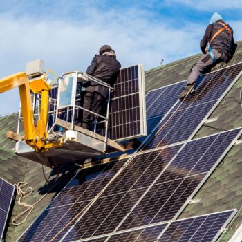 Workers installing solar panels on private home hexagonal roof felt on sunny day, blue sky. Photo via Adobe Stock.