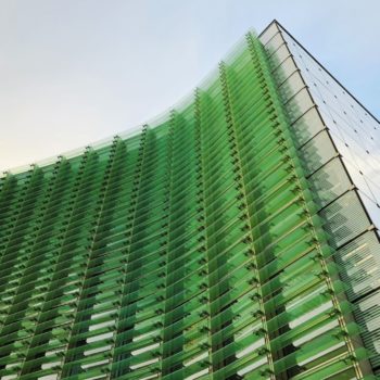 Upward-facing shot of the National Art Center in Tokyo, Japan. This shows the exterior of the curved glass building, tinted green, against a bright blue sky and white clouds. The shape resembles a cresting wave.