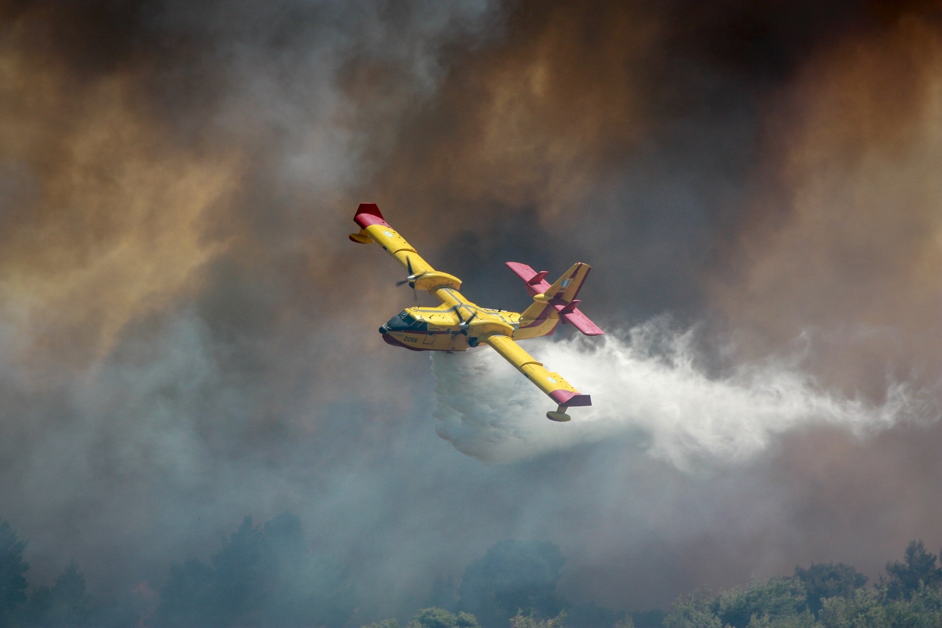 The image shows a plane, a North American P-51 Mustang, fighting a wildfire in Varnavas, Greece. There is a lot of smoke and water coming out of the plane.